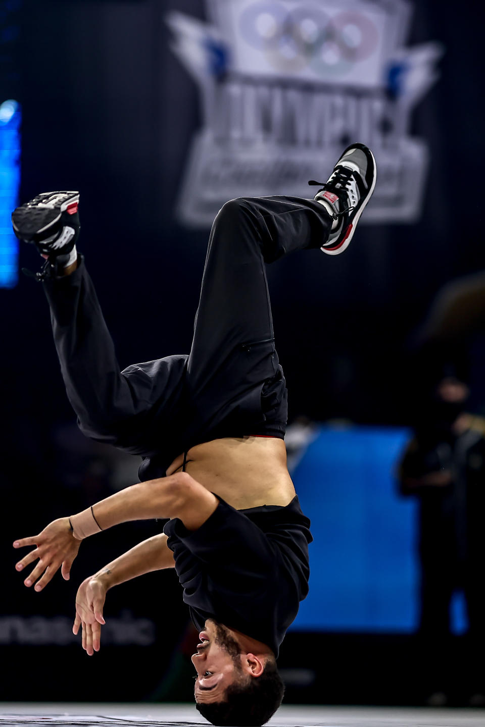 A breakdancer performs an inverted headspin on stage during an event with the Olympic rings symbol visible in the background