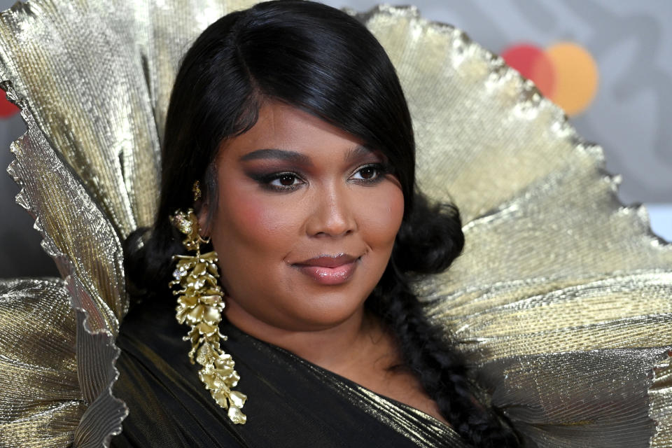 Lizzo on the red carpet, wearing an elaborate gold and black gown with dramatic ruffled shoulders and gold leaf earrings