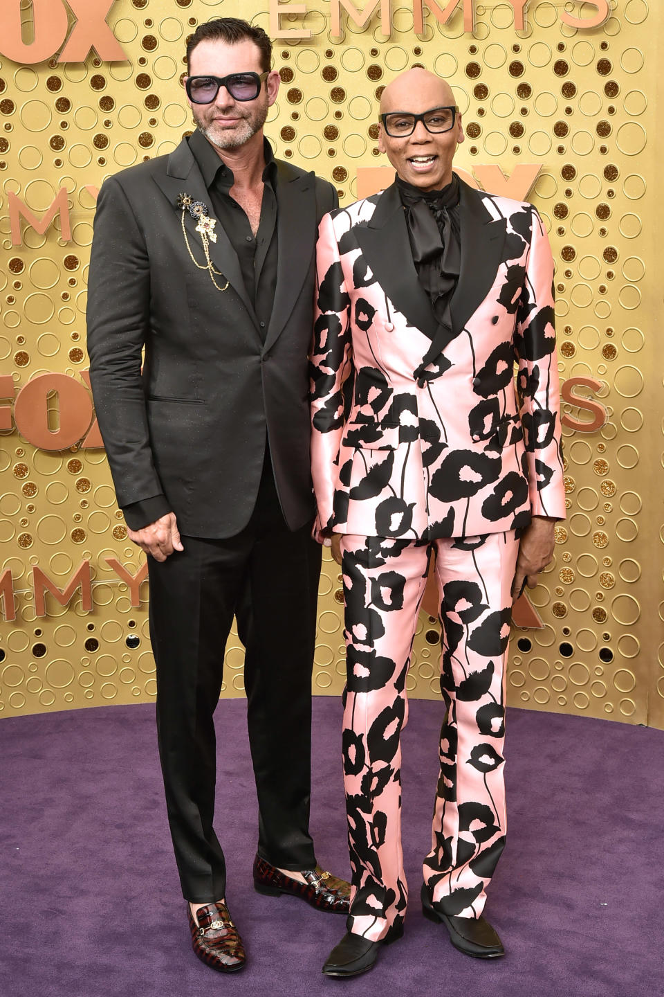 RuPaul is wearing a floral-patterned suit and black blouse, standing next to Georges LeBar in a dark suit with a brooch on a red carpet