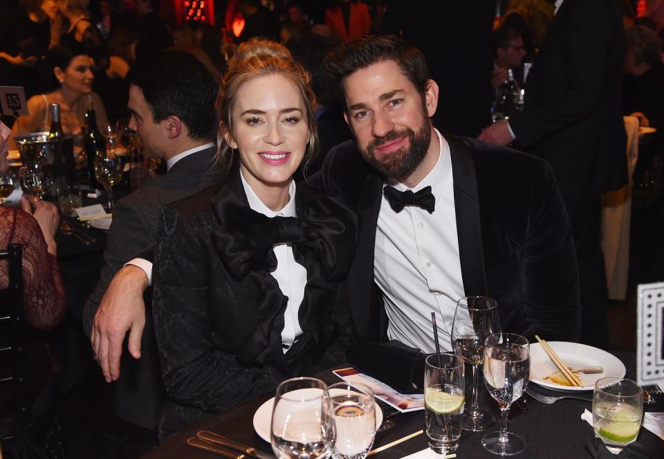 emily blunt and john krasinski sit at a table and smile for a photo, both wear black bow ties with white collared shirts, she also wears a black jacket, he has his arm around the back of her chair