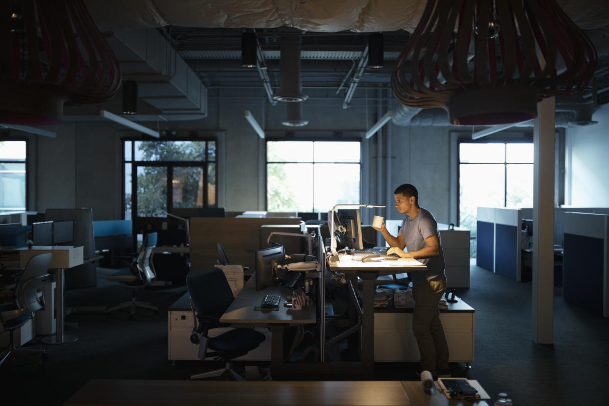 Dedicated businessman working late at computer, drinking coffee at sit-stand desk in dark office