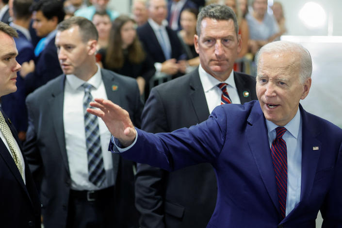 Biden reaches out with his hand to wave, with bodyguards behind him.