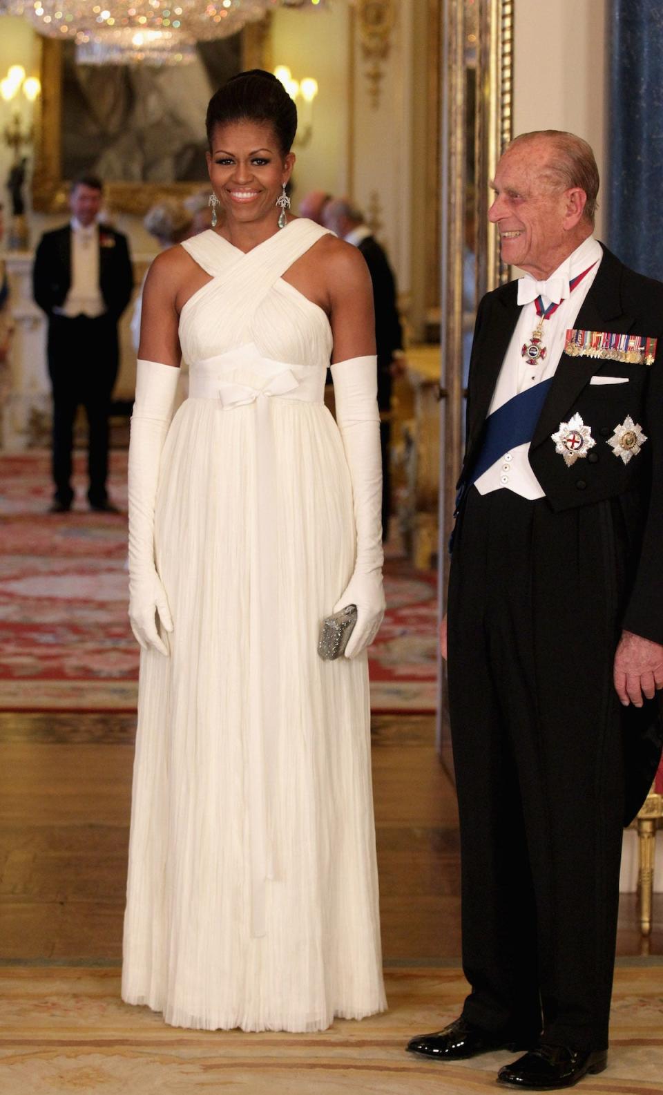 Michelle Obama wears a white dress and stands next to Prince Phillip.