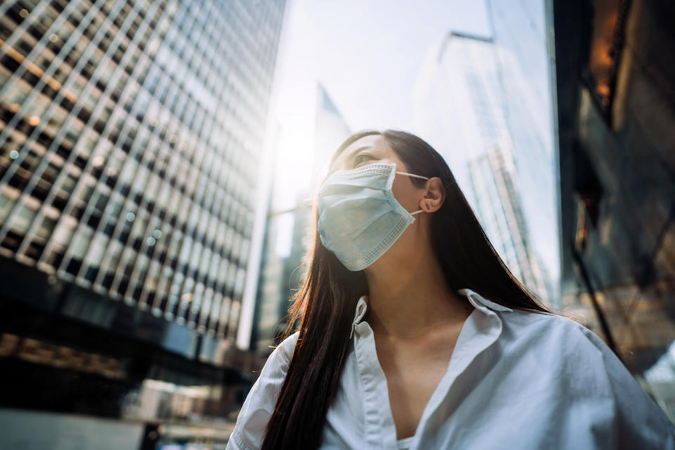 Young Asian businesswoman with protective face mask to protect and prevent from the spread of viruses during the coronavirus health crisis, standing in an energetic and prosperous downtown city street against corporate skyscrapers