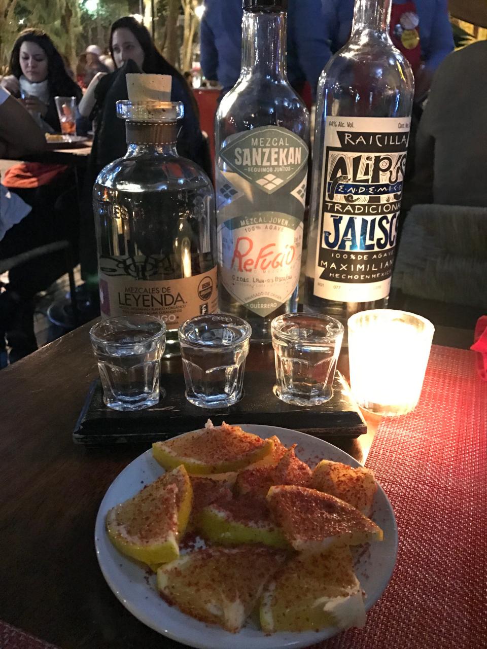 Three bottles of Mezcal and three shot glasses on a table with a bowl of lemon wedges in CDMX