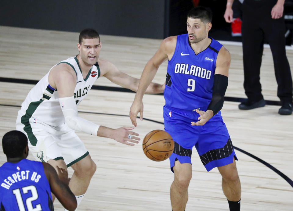 Orlando Magic center Nikola Vucevic (9) passes the ball to forward Gary Clark (12) in front of Milwaukee Bucks center Brook Lopez (11) during the first half of Game 1 of an NBA basketball first-round playoff series, Tuesday, Aug. 18, 2020, in Lake Buena Vista, Fla. (Kim Klement/Pool Photo via AP)