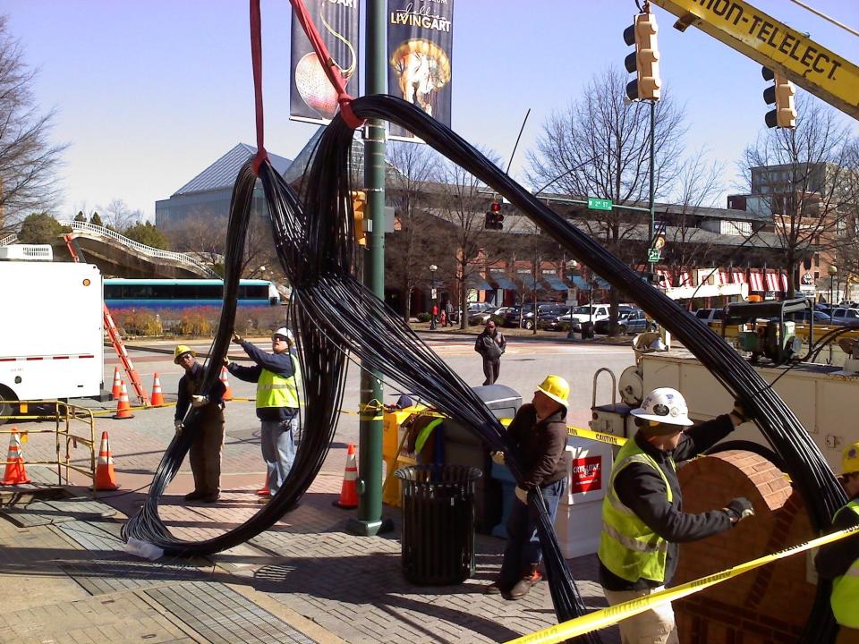 utility workers in hard hats handle thick bundles of fiber optic cable which a vehicle arm is lifting upward toward power lines