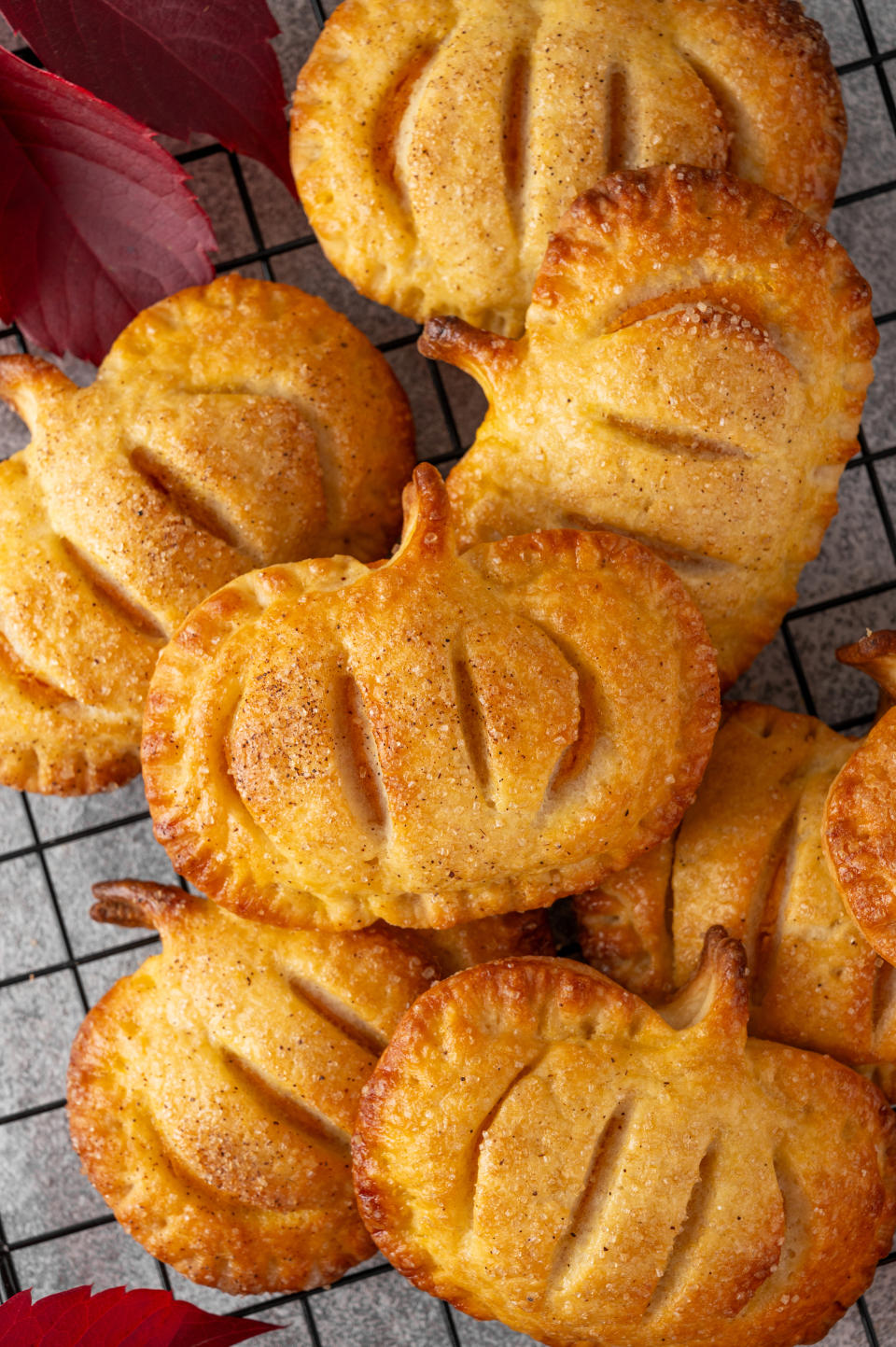 A batch of pumpkin-shaped pastries with slits on top, cooling on a wire rack next to autumnal leaves