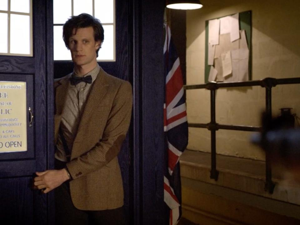 Matt Smith walking out a blue police box in a brown blazer and black bow tie for "Doctor Who."