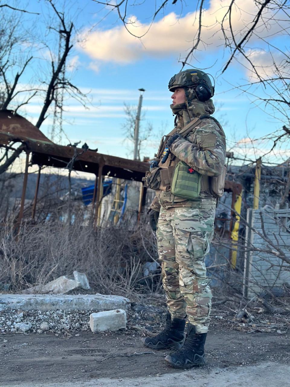 Katerina Zirka stands in combat gear in front of rubble and a blue sky