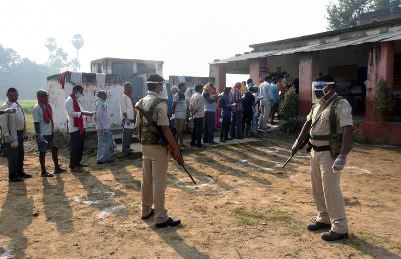 Policemen wearing protective face shields stand guard as voters stand in a queue to cast their vote outside a polling booth during the state assembly election, at a village on the outskirts of Patna