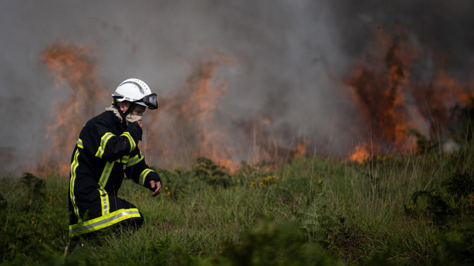 A firefighter covers their mouth with a cloth as they walk near smoke and flames burning in vegetation.