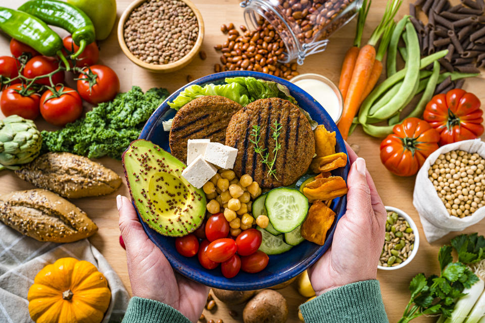 Overhead view of woman's hands holding a plate filled with healthy plant-based food. The composition includes soy beans, soy milk, tofu, legumes, leaf vegetables, root vegetables, wholegrain pasta, avocado, chia seeds and veggy patties. High resolution 42Mp studio digital capture taken with SONY A7rII and Zeiss Batis 40mm F2.0 CF lens