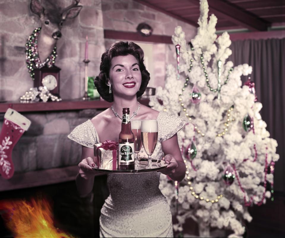 Portrait of an unidentified model as she stands in front of a Christmas tree next to a fireplace with a bottle of Pabst Blue Ribbon beer, two glasses, and a present on a serving tray, 1951. (Photo by Tom Kelley/Getty Images)