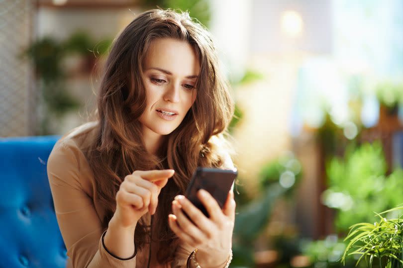 Green Home. stylish woman with long wavy hair in the modern house in sunny day sending text message using smartphone.