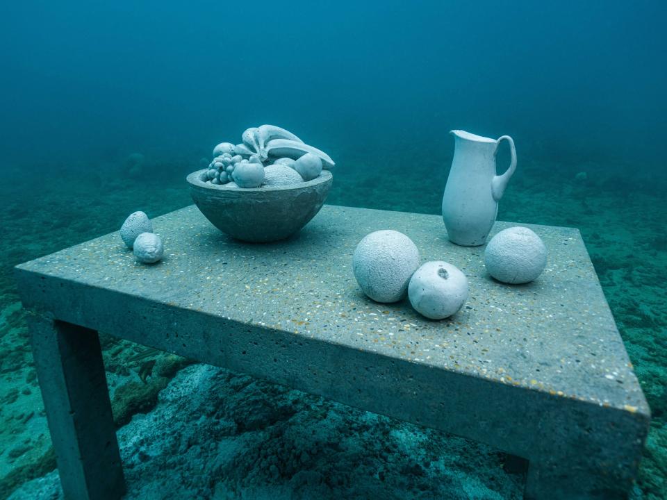An underwater sculpture of a table with a bowl of fruit, a pitcher, and loose fruit.