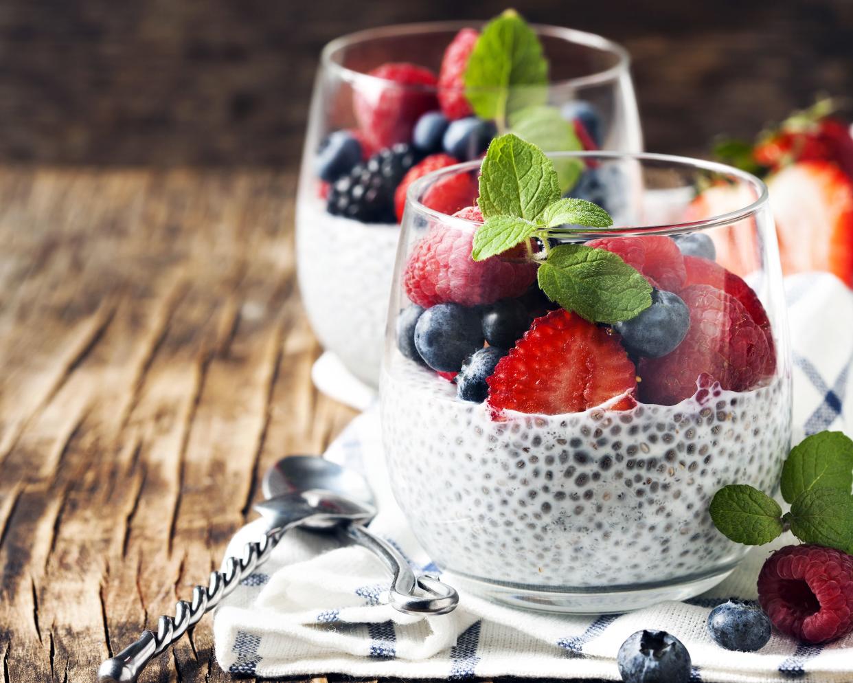 Two basic chia seed pudding in glass cups with strawberries, raspberries, blueberries, and blackberries, with two spoons on a blue-lined white napkin, on the right, on a rustic wooden bench with a blurred background
