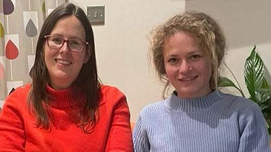 Two women, Sarah and Yulia, sit next to each other and smile at the camera