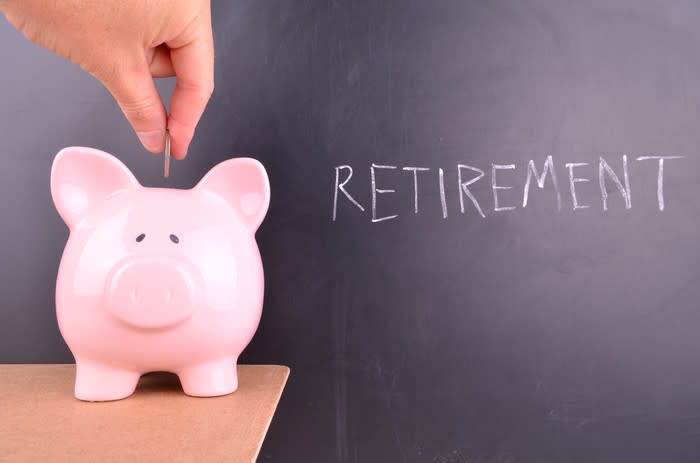 Person inserting coin into piggy bank sitting on a table in front of a blackboard with the word retirement written on it in chalk