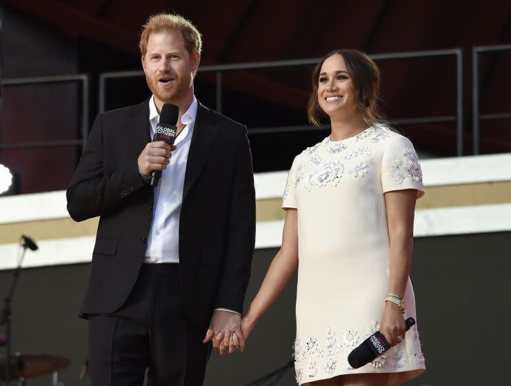 Prince Harry in a black blazer and pants standing next to and holding hands with Meghan Markle in a white dress