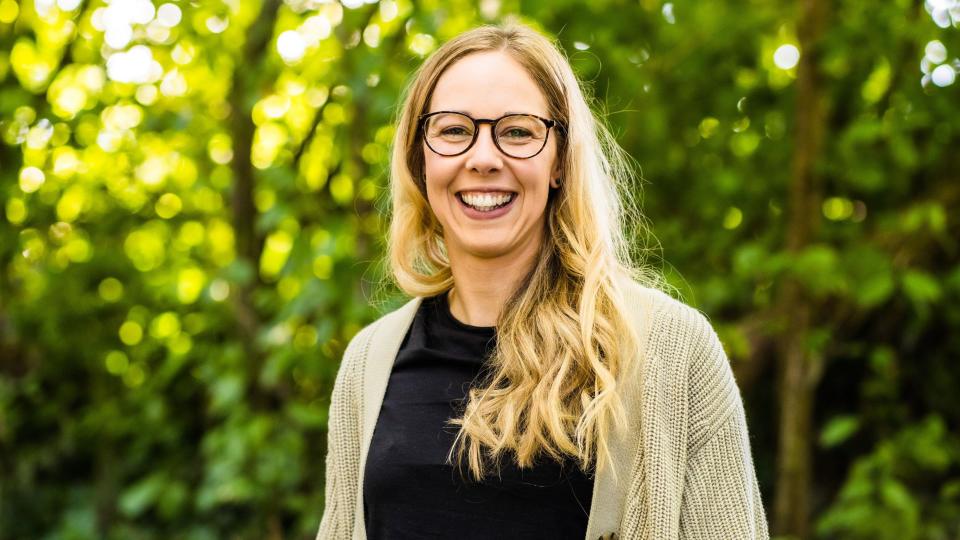 A blonde woman with long hair over her shoulder, smiles at the camera wearing round glasses in front of some trees