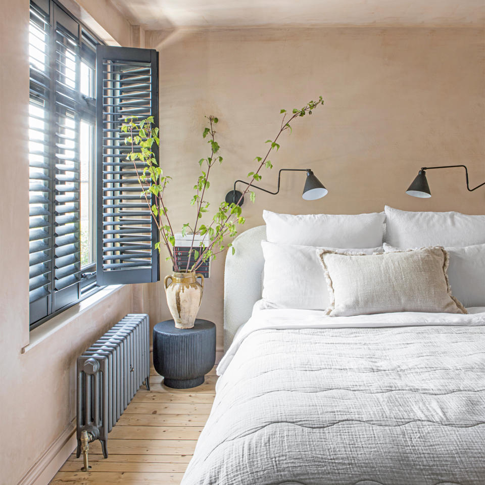 A bedroom with bare plaster walls and navy blue shutters on the windows with a bed dressed in white bedlinen