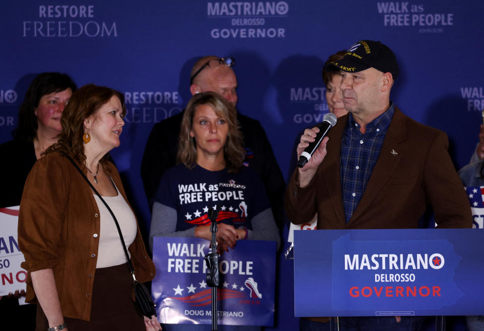 Doug Mastriano, in U.S. Army baseball cap, speaks onstage, as his wife, Rebbie, looks at him with a sympathetic expression.