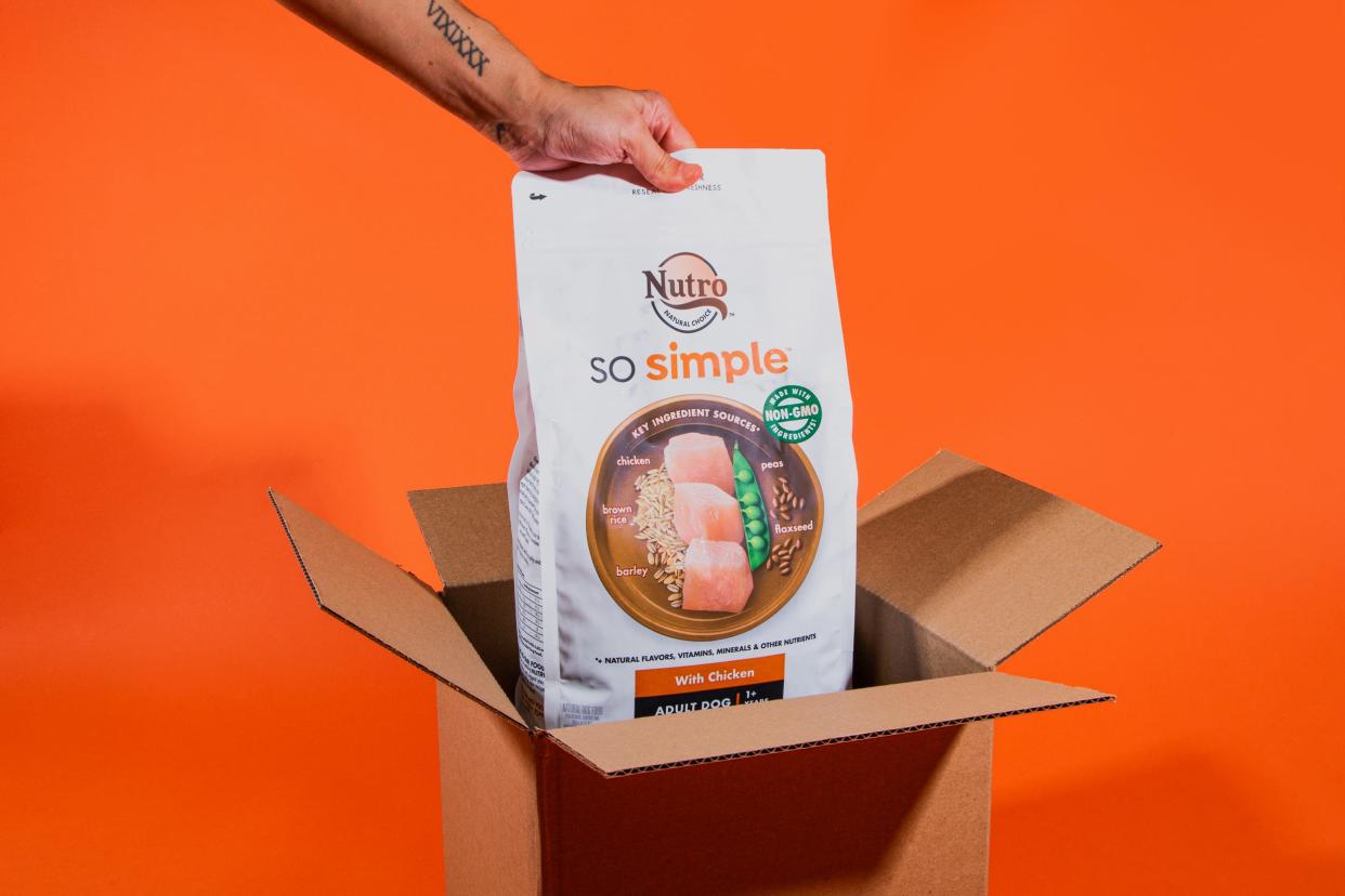 A hand pulls a bag of dog food out of a cardboard box, on an orange background.
