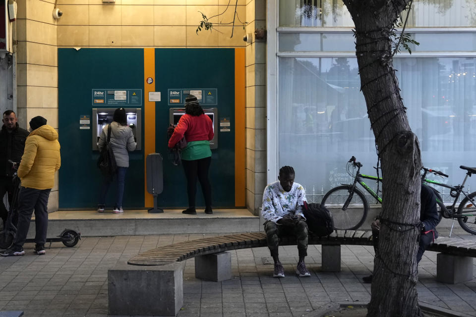 A migrant sits on a bench as others use an ATM machine at Elepfhteria, Liberty Square in divided capital Nicosia, Cyprus, Saturday, Jan. 28, 2023. Cypriots are voting Sunday for a new president who they’ll expect to decisively steer the small island nation through shifting geopolitical sands and uncertain economic times that have become people's overriding concern, eclipsing stalemated efforts to remedy the country’s ethnic division. (AP Photo/Petros Karadjias)