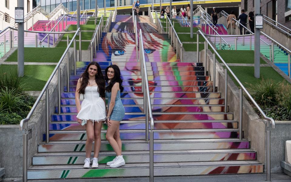 Two Taylor Swift fans pose in front of a mural depicting the singer on the steps of Wembley Stadium