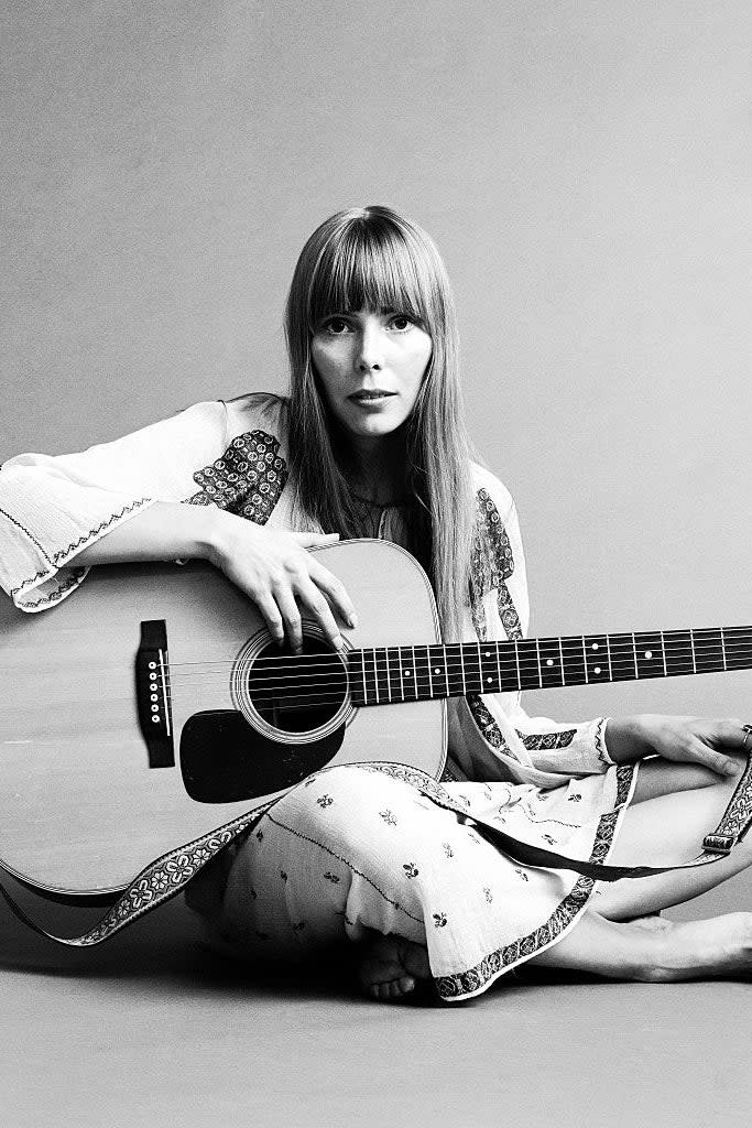 Joni Mitchell sits barefoot, holding an acoustic guitar in a folk-style dress with intricate patterns