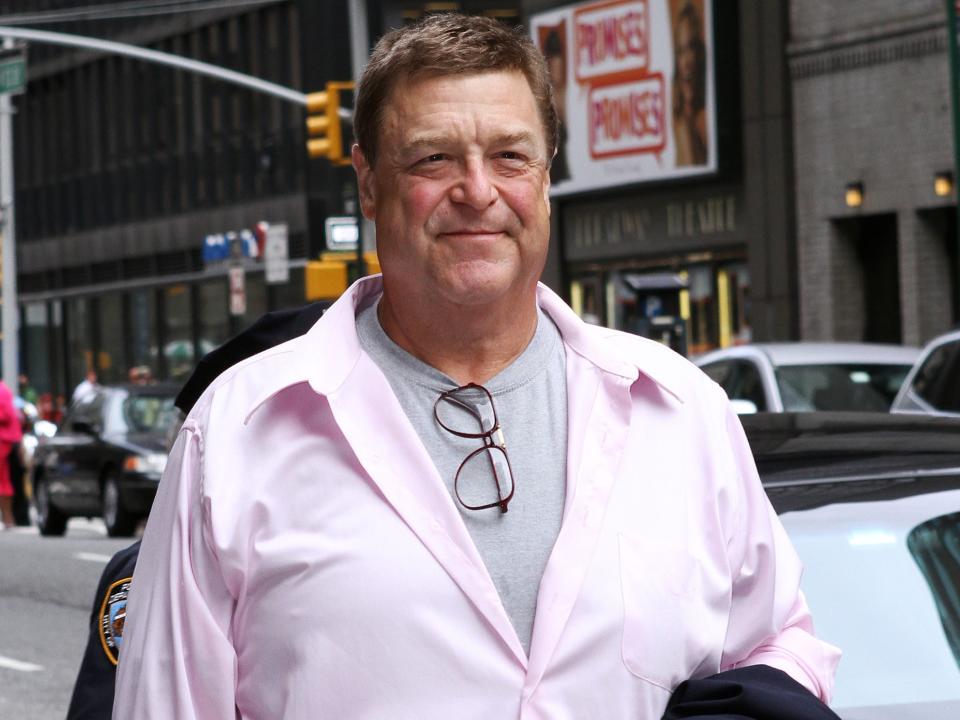 Goodman wearing a light pink shirt and a grey under shirt while standing on a busy sidewalk.