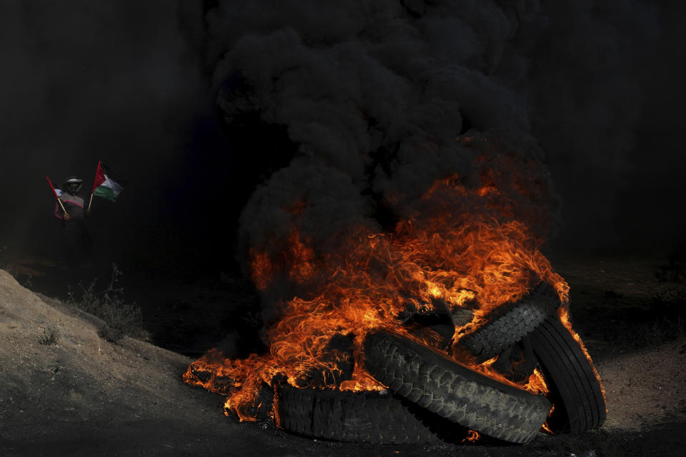 A Palestinian demonstrator waves two Palestinian flags while others burn tires during a protest against an Israeli military raid in the West Bank city of Jenin, along the border fence with Israel, east of Gaza City, Monday, July 3, 2023. Israel struck targets in a militant stronghold in the occupied West Bank with drones early Monday and deployed hundreds of troops in the area. Palestinian health officials said at least eight Palestinians were killed. (AP Photo/Adel Hana)