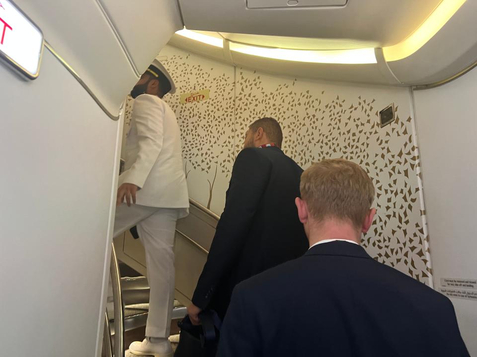 Three men are viewed from behind ascending the winding staircase of Emirates Airbus A380, with an intricate gold design on the white wall