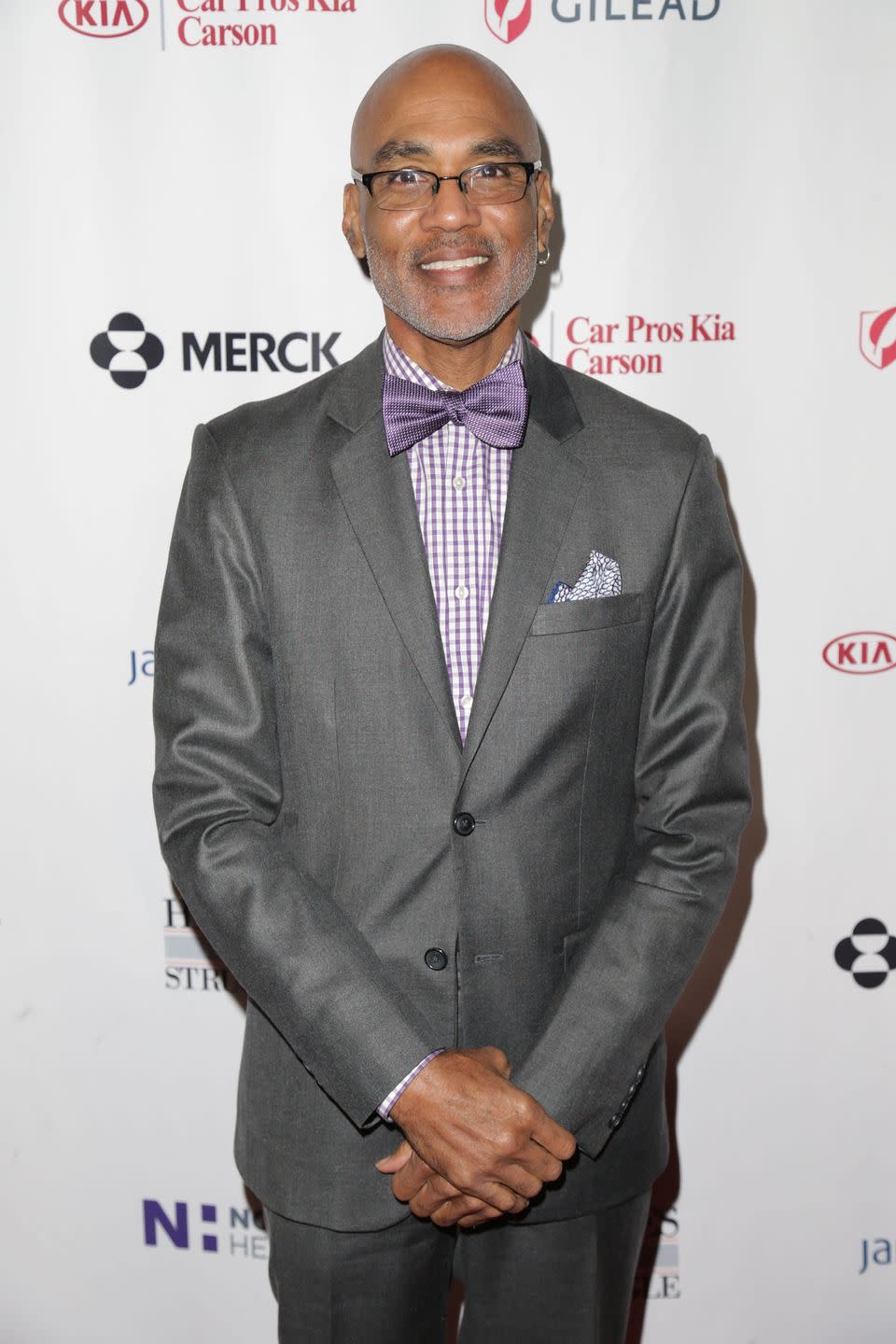 phil wilson smiles at the camera while standing in front of a white background with logos, he clasps his hands in front of him, he wears a gray suit with a purple and white checkered shirt and purple bowtie