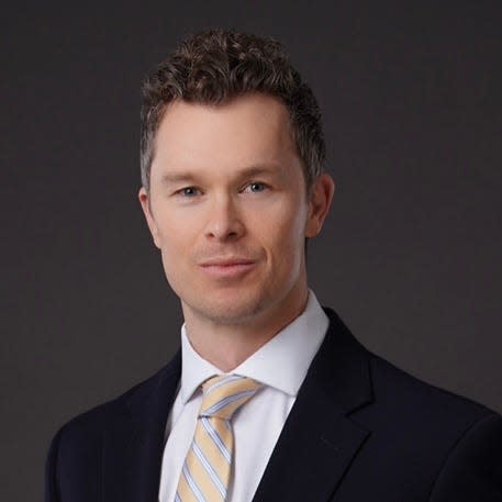 A picture of a man with brownish short curly hair in a black jacket and a tie with blue and orange stripes.