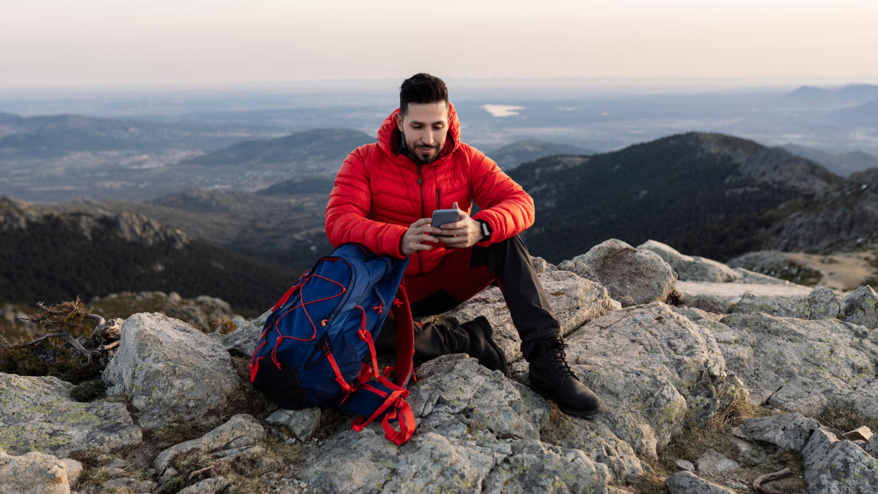  Man sitting on rocky mountain checking smartphone. 