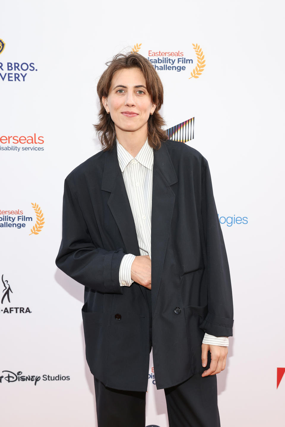 Rhea Butcher at the Easterseals Disability Film Challenge event, wearing an oversized black blazer over a white-striped shirt, standing in front of a step-and-repeat backdrop