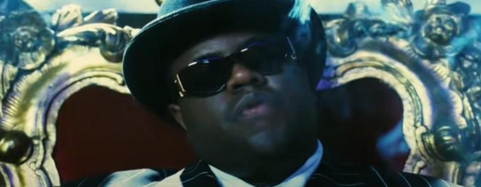 Jamal Woolard as Biggie, or the Notorious B.I.G., leaning back with a black bowler hat and sunglasses on, seated in a red velvet chair with ornate gold edges