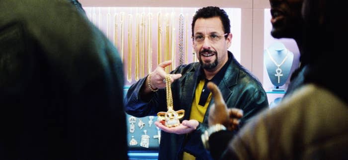 Adam Sandler wearing a leather jacket, holding a gold chain with a pendant shaped like a furby, in a jewelry store display setting