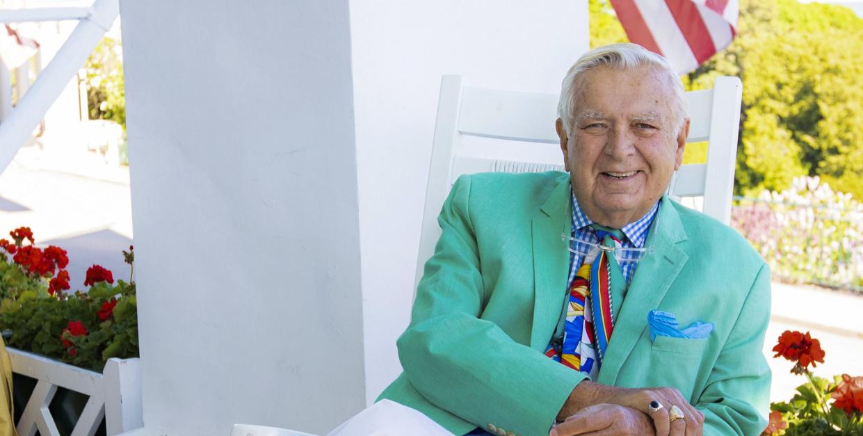 a man in a green jacket and white pants sits on a white rocking chair on a porch with an american flag in the background