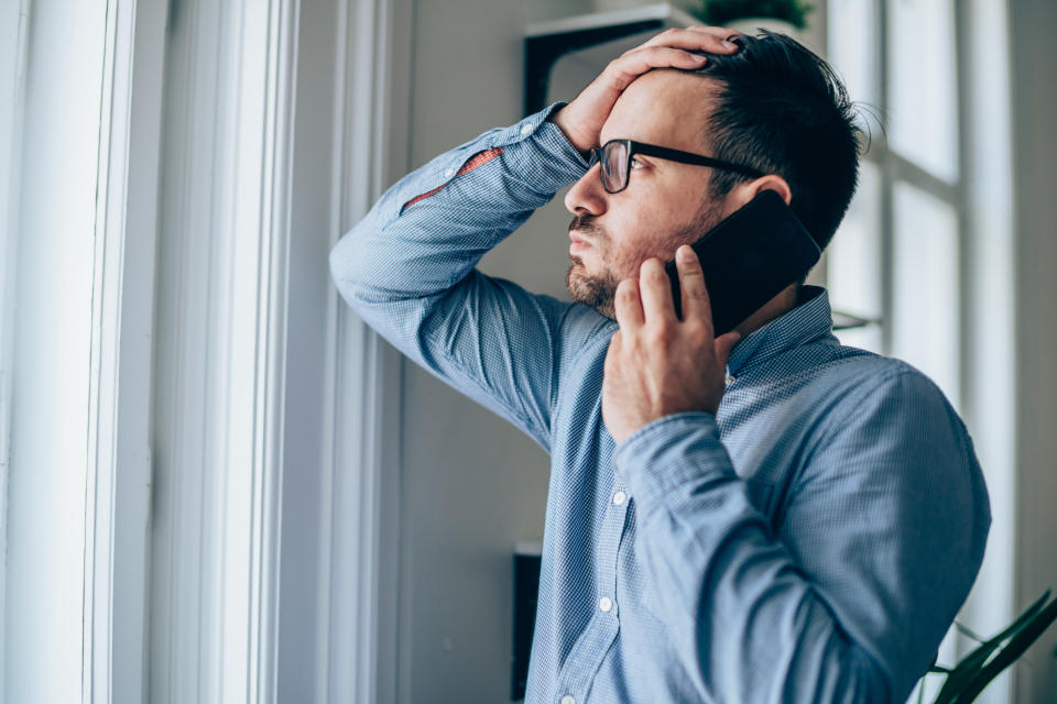 A man in a button-up shirt and glasses talks on his phone while looking out a window, his other hand resting on his head in a contemplative gesture