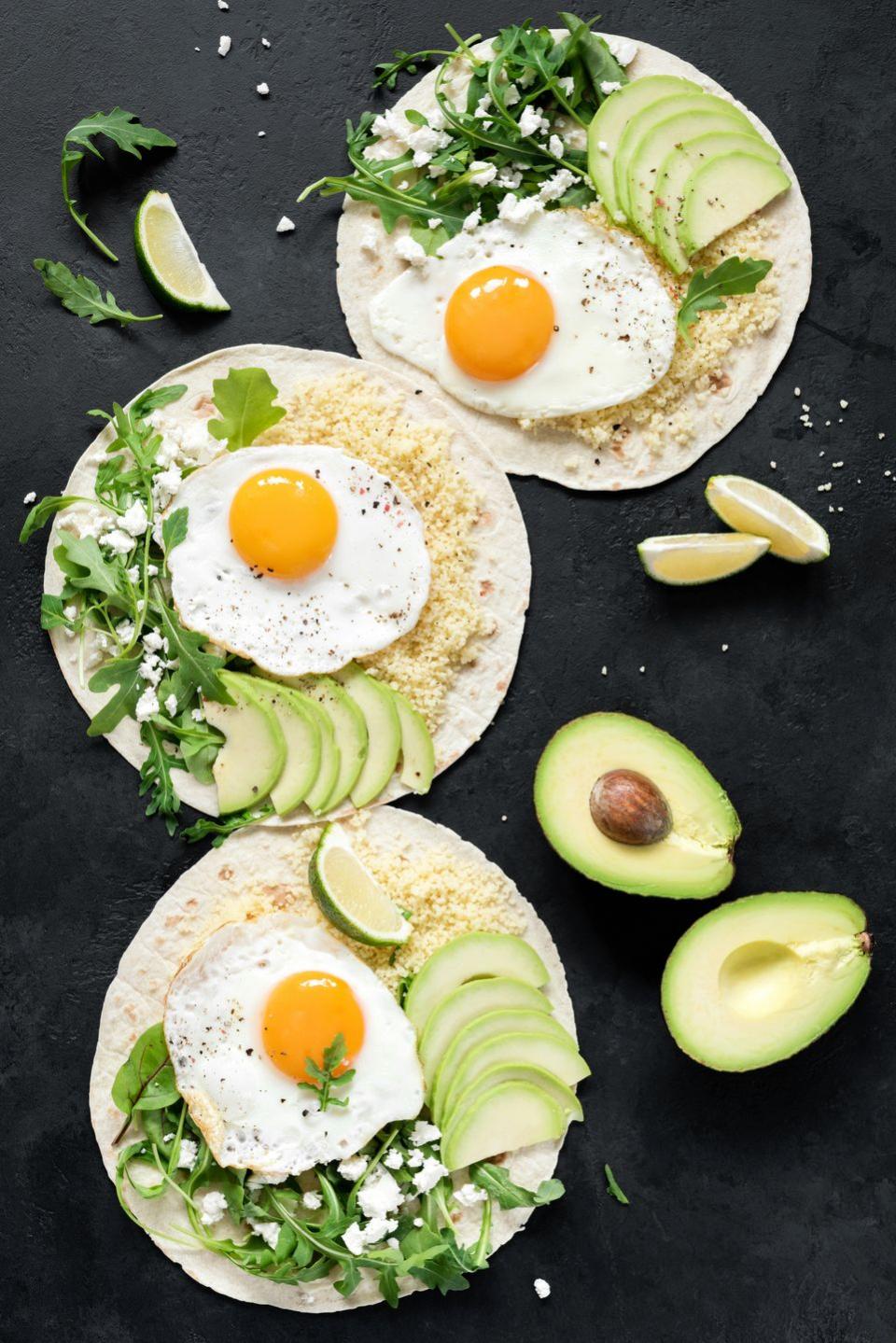 healthy tortilla or flatbread with sliced avocado, fried egg, couscous and arugula on black slate background