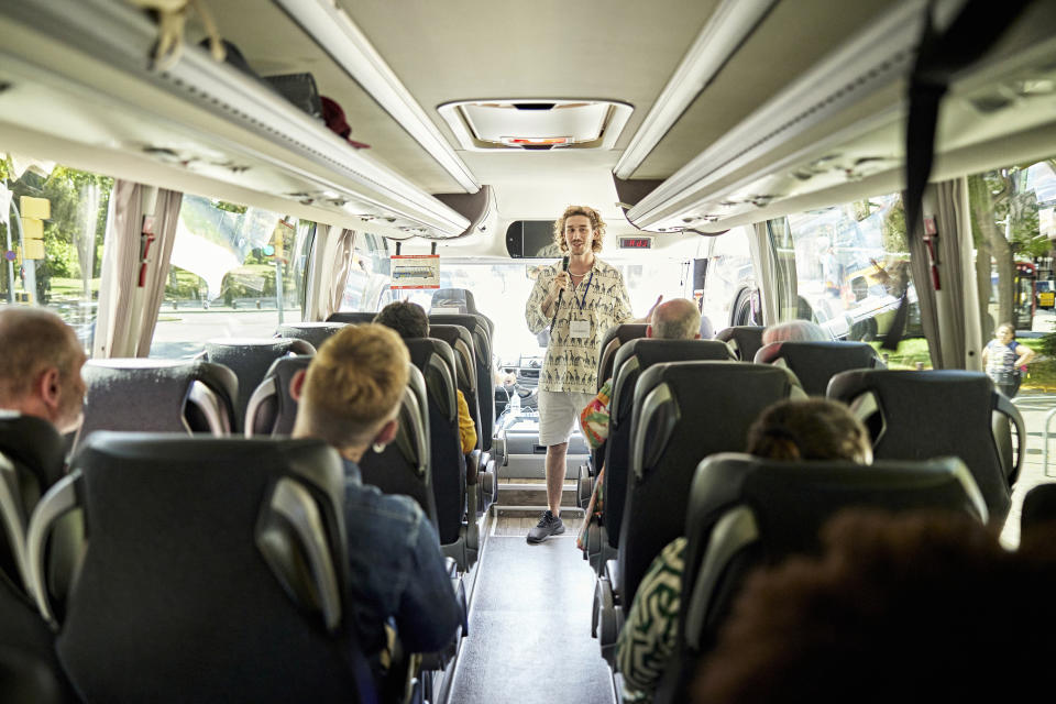 Tour guide standing in the aisle of a bus, speaking to passengers, wearing casual attire