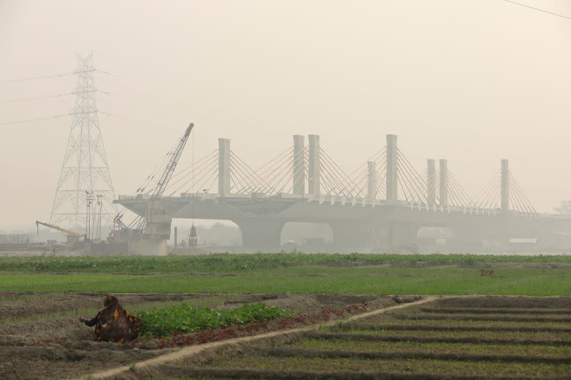 A woman works on the fields next to a construction site of a bridge on the field on the Yamuna floodplains on a smoggy day in New Delhi