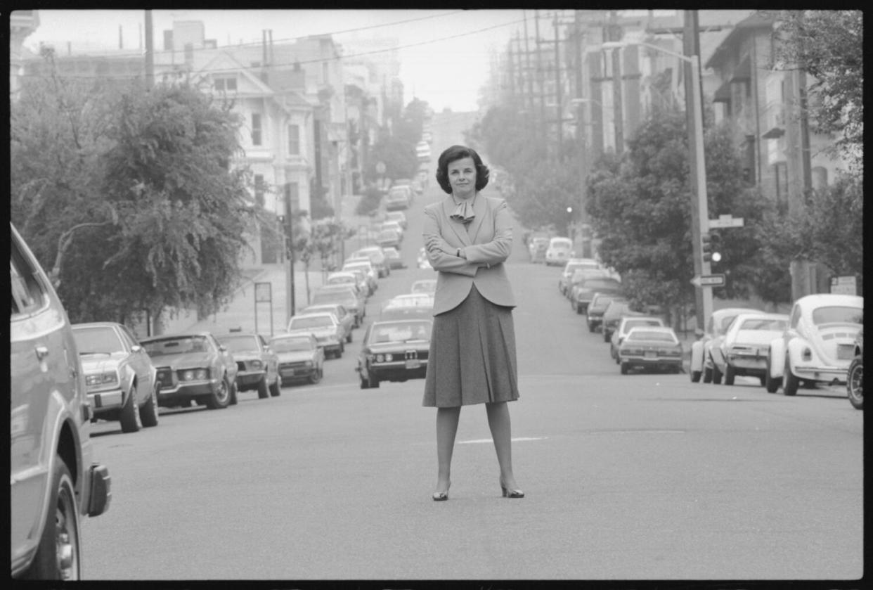 A black-and-white photo of Dianne Feinstein standing in the middle of a street with her arms crossed.