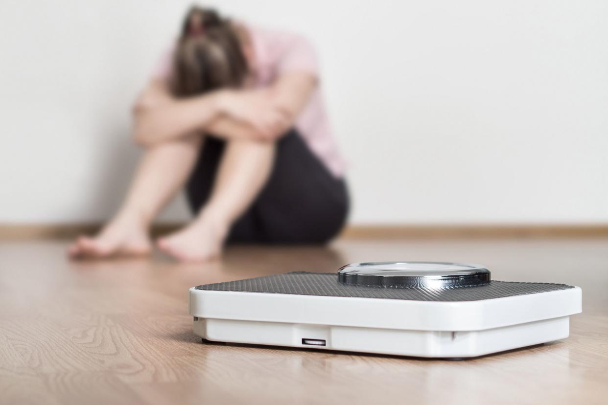 A weight scale in the foreground, selective focus, on the floor, an upset woman sitting on the floor with her head hidden behind her arms on her knees, blurred, while sitting against a white wall