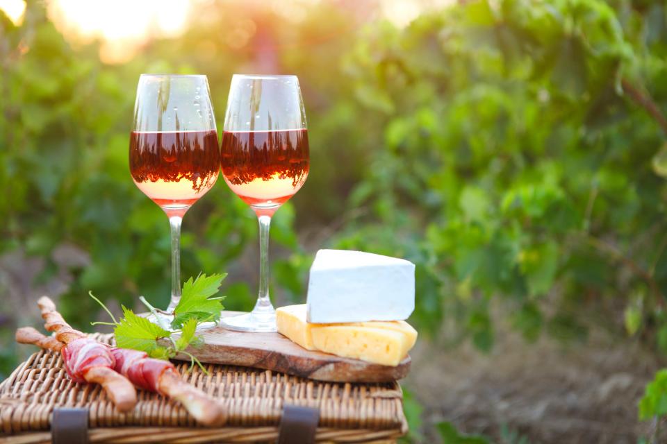 Two glasses of rose wine with bread, meat, grape and cheese laid out on a picnic basket. Rose wine is ideal for a picnic setting.
