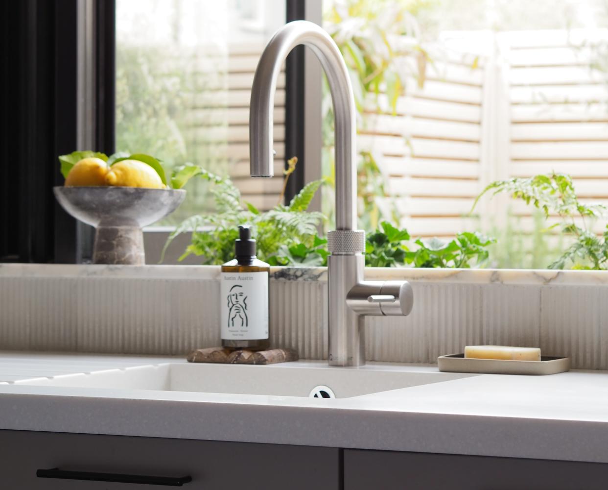  A kitchen sink with a brushed steel faucet, a white quartz countertop, a bar of soap, and a window with a view into a garden. 