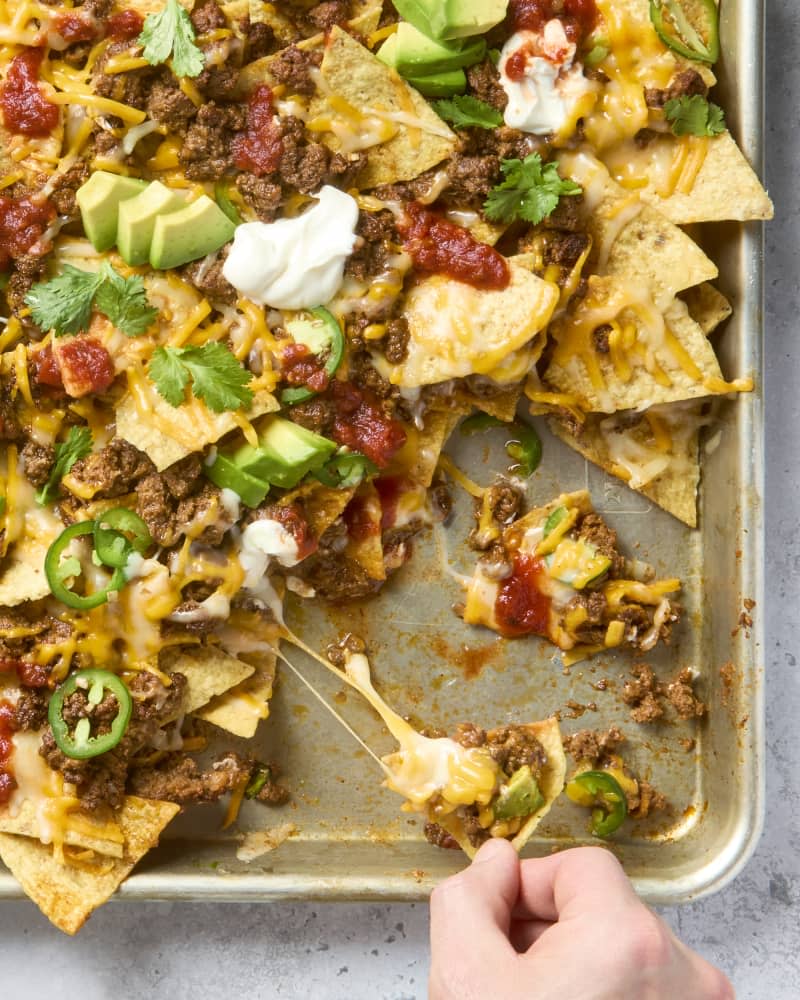 overhead shot of nachos in a sheet pan, with some taken from the bottom right corner, topped with cilantro, avocado, sour cream and salsa.